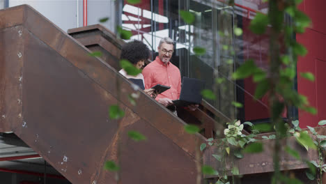 Diverse-male-and-female-colleague-walking-down-stairs-talking,-using-tablet-and-laptop,-slow-motion