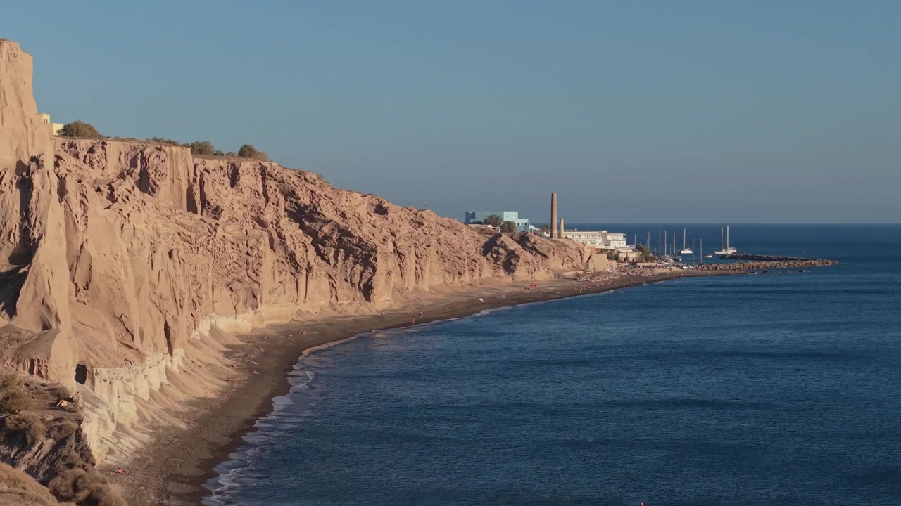 Luftfotoafsløring af Vlychada Strand, der viser dramatiske vulkanske formationer, den klippefyldte kystlinje og den charmerende havn sat mod Santorinis landskab