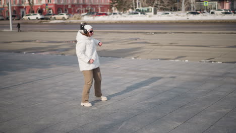 young woman wearing white winter coat, reflective glasses, and headphones dances joyfully on tiled plaza in bright light, bordered by snow piles beside busy road under clear blue winter sky