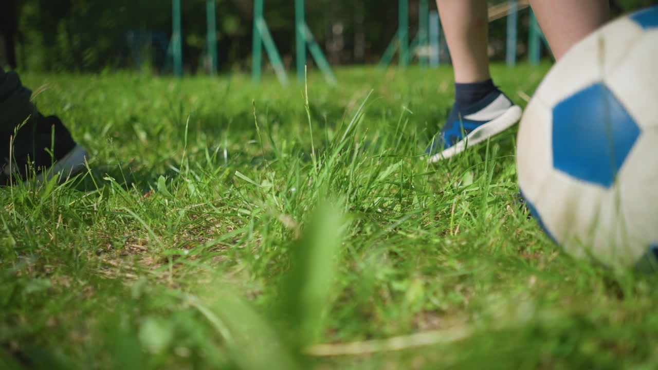 близкий вид маленького ребенка на травяном поле, с футбольным мячом поблизости, на заднем плане, другие могут быть замечены прыгающими, а на заднем фоне видно оборудование для отдыха