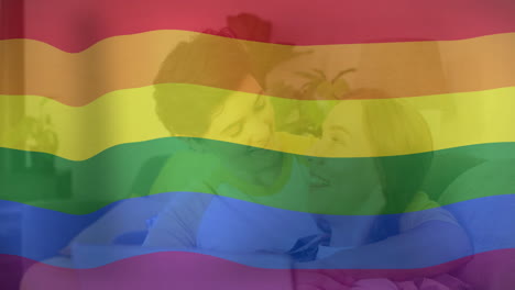 animación de la bandera del arco iris del orgullo sobre una feliz pareja de lesbianas caucásicas usando la computadora portátil en la cama