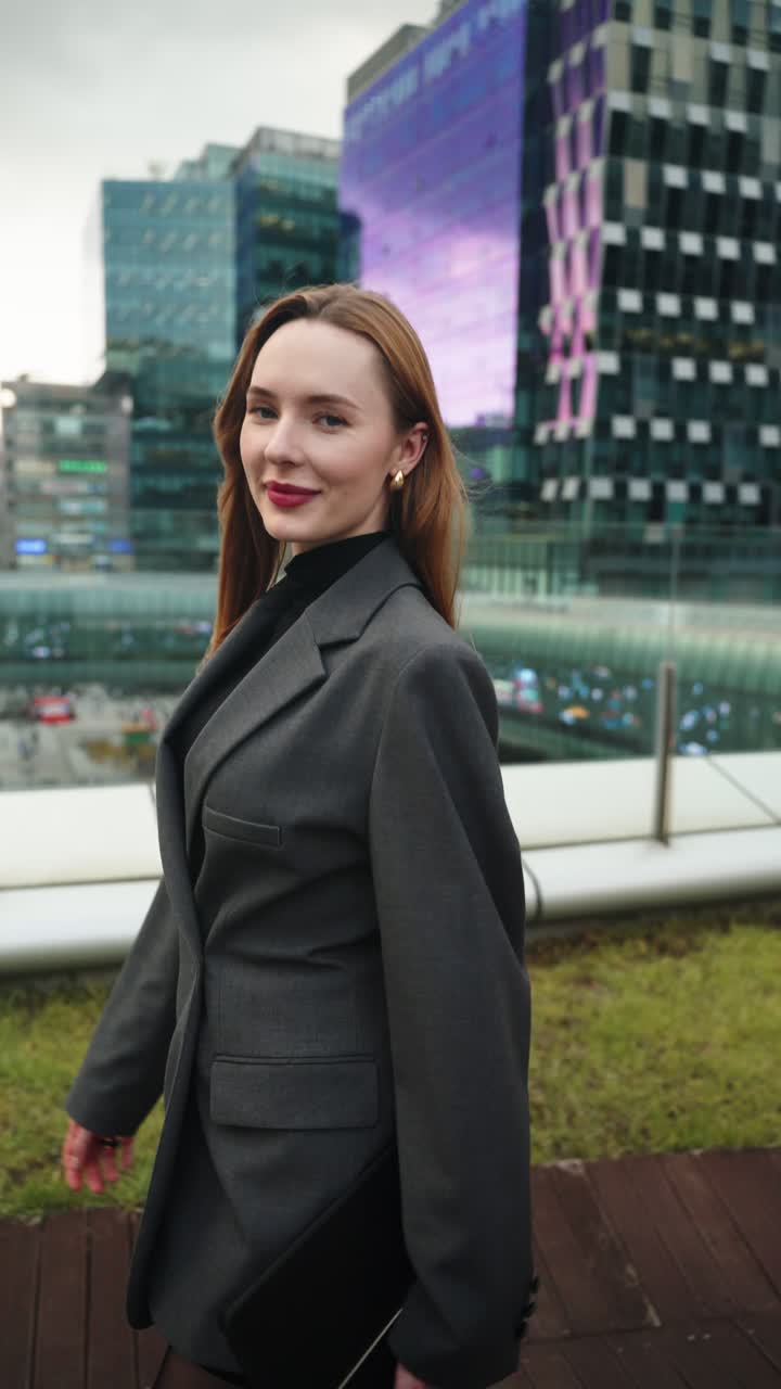 Confident businesswoman in gray blazer and tights walks on rooftop terrace holding tablet and smartphone in urban business district