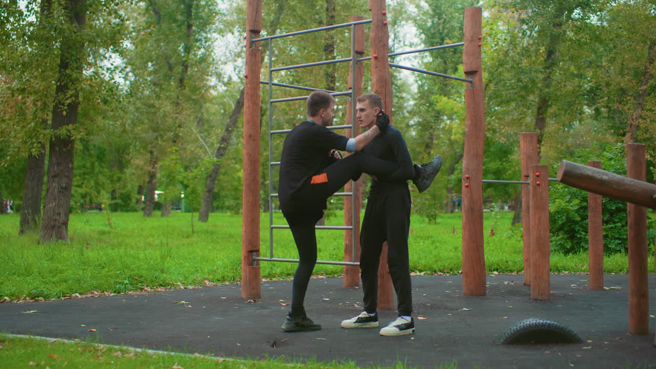 white male instructor teaching kick then takedown at calisthenics park, wooden posts and ladder backdrop, partner receives strike then rolls to ground, rubber surface, dynamic fall and ground control