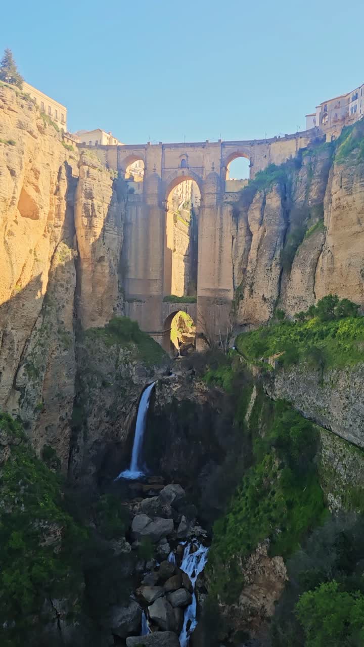 Una vista dramática del histórico puente de Ronda que atraviesa un cañón profundo con una cascada fluyendo debajo. Acantilados iluminados por el sol y exuberante vegetación enmarcan la escena.