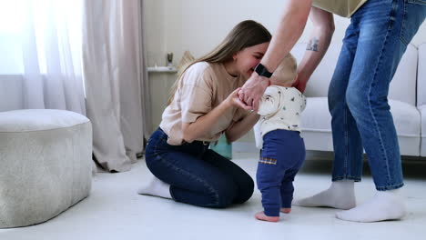 unrecognized man wearing jeans holds the stepping kid by the hands. mother sitting on the floor calls up her son.