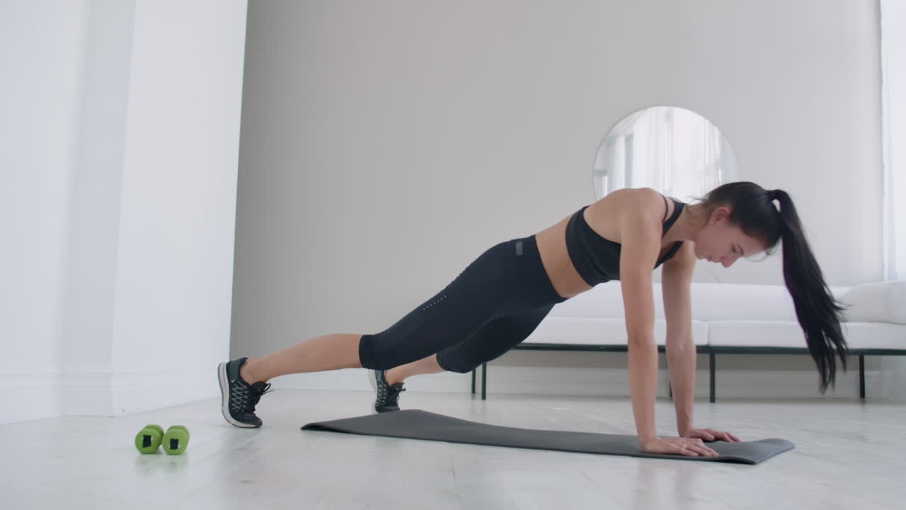 A young brunette in black clothes and a light apartment performs the plank exercise jumping in the rest lying down