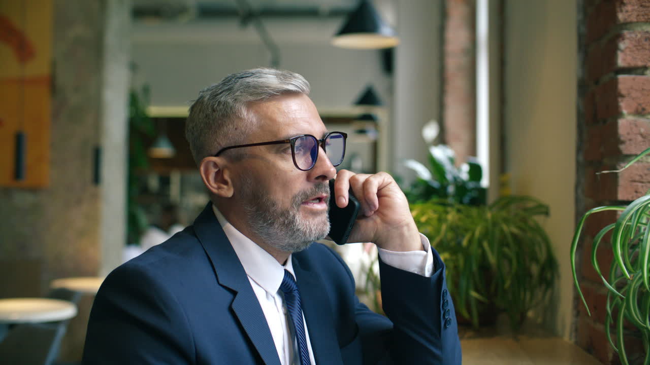 Imprenditore senior seduto vicino alla finestra in un bar, impegnato in una conversazione telefonica