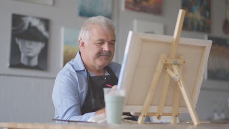 an elderly man in an apron paints a picture with a brush while sitting at the table