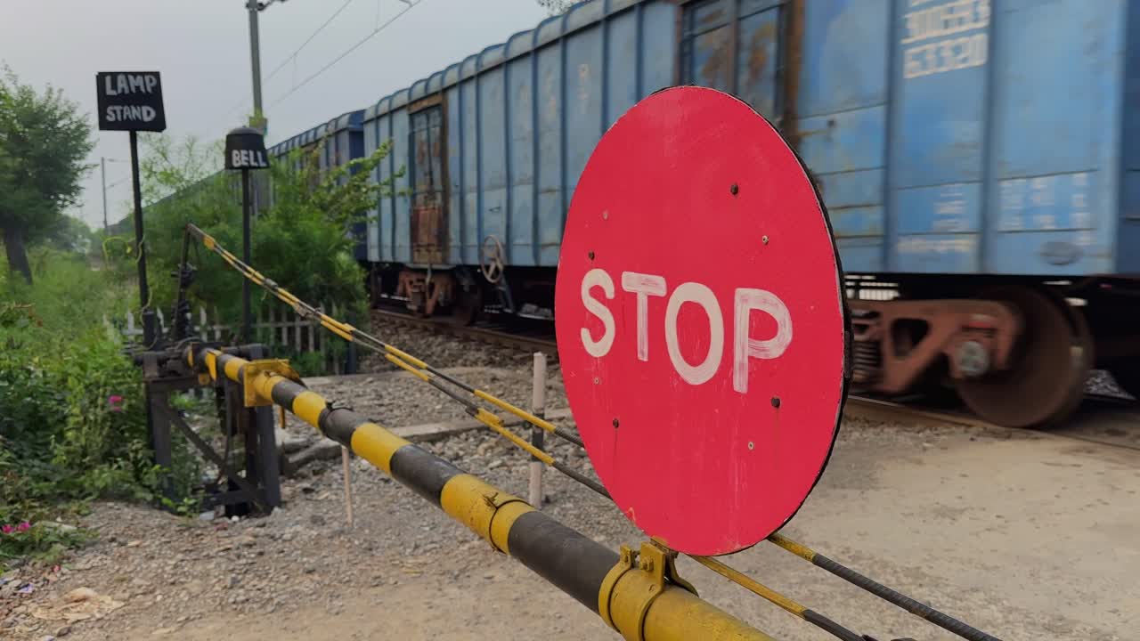 Freight train moves past a railway crossing while a red stop sign stands ahead, with yellow-black barrier poles, gravel ground, metal tracks, and soft daylight shaping a moving roadside scene