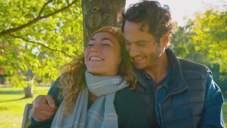 Happy-Loving-Couple-Smiling-As-They-Sit-On-Bench-In-Autumn-Park-And-Hug