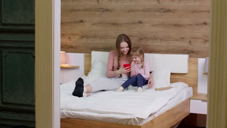 madre acostada en la manta de la cama con la hija niña con teléfono móvil viendo dibujos animados