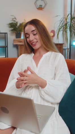 happy young woman making online video call on laptop talking waving hand hello hi sitting on sofa