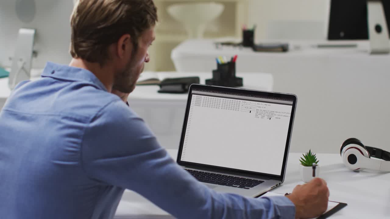 hombre caucásico sentado en un escritorio viendo el procesamiento de datos de codificación en una computadora portátil y tomando notas
