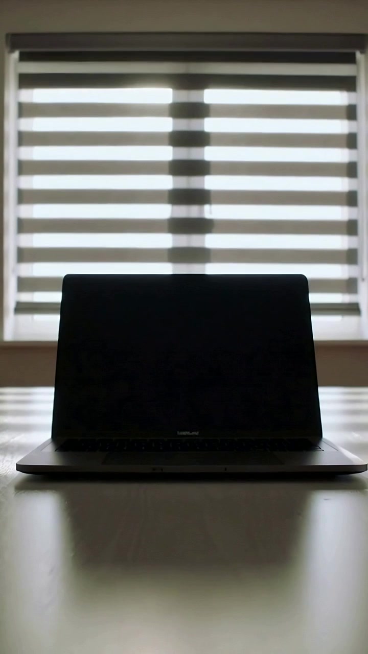 Vertical video: Camera zooming in, dark laptop growing on table at home near blinds
