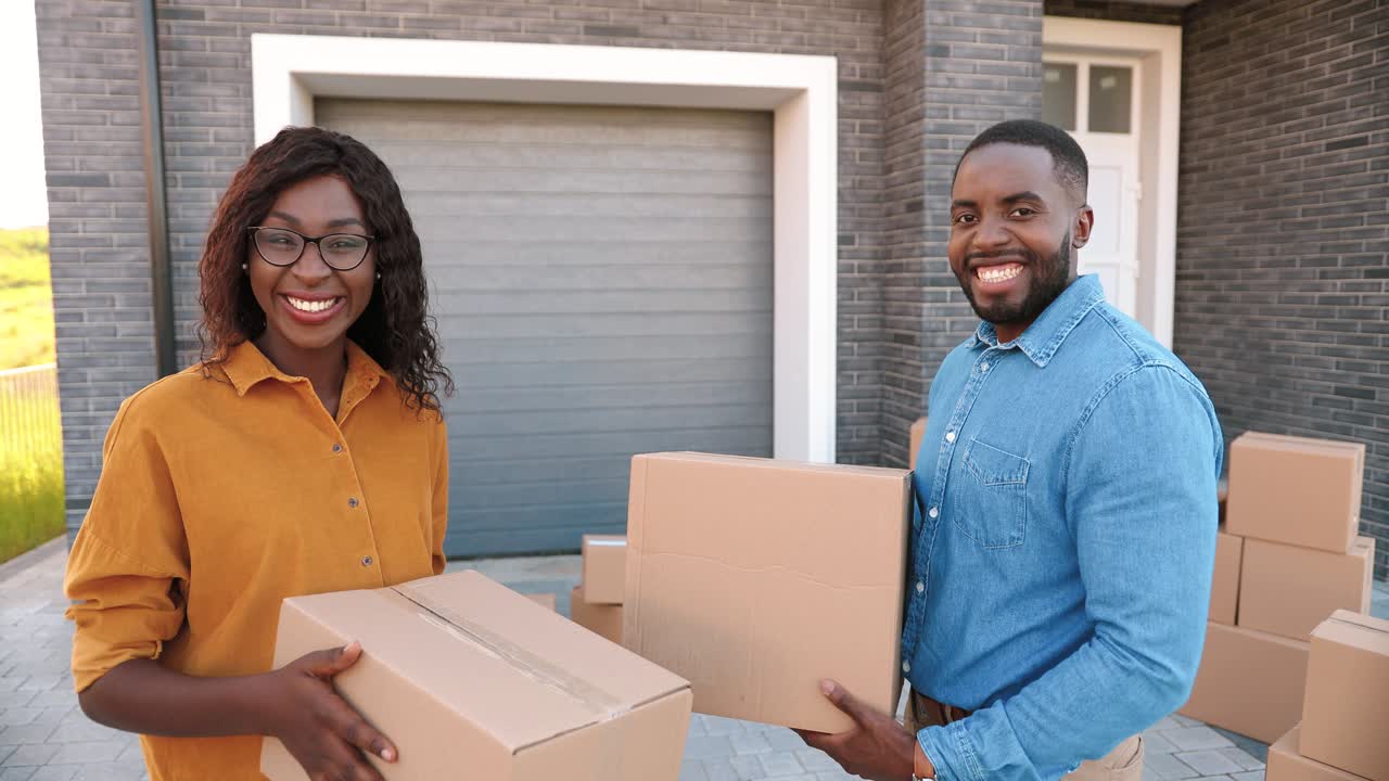 Rear view on African American young couple walking in yard at big house and carrying carton boxes