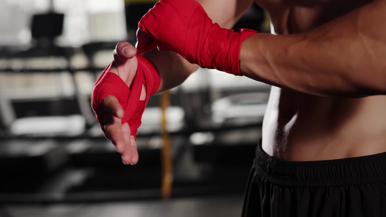 Ein Boxer bereitet sich auf Training, Fitness oder einen Kampf vor, indem er rote Bandagen um seine Hände wickelt, vor einem Boxring, was Fitness und einen aktiven Lebensstil im Fitnessstudio unterstreicht.