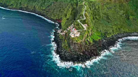 A-lighthouse-perched-on-a-rugged-green-cliff-by-the-ocean,-aerial-view