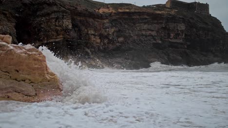 SLOW-MOTION-waves-white-with-foam-break-on-a-sandy-rocky-cliff-with-old-castle-on-the-dark-mountains-in-the-background,-Cape-Roca-Portugal