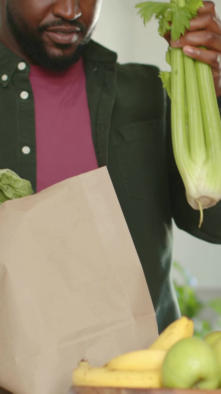 Video vertical: Hombre con camisa verde colocando una bolsa de papel marrón en la encimera de la cocina, sacando apio y desempacando.