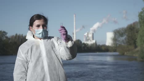 woman in face mask and protective suit holding test tube with water sample, smoking factory in background