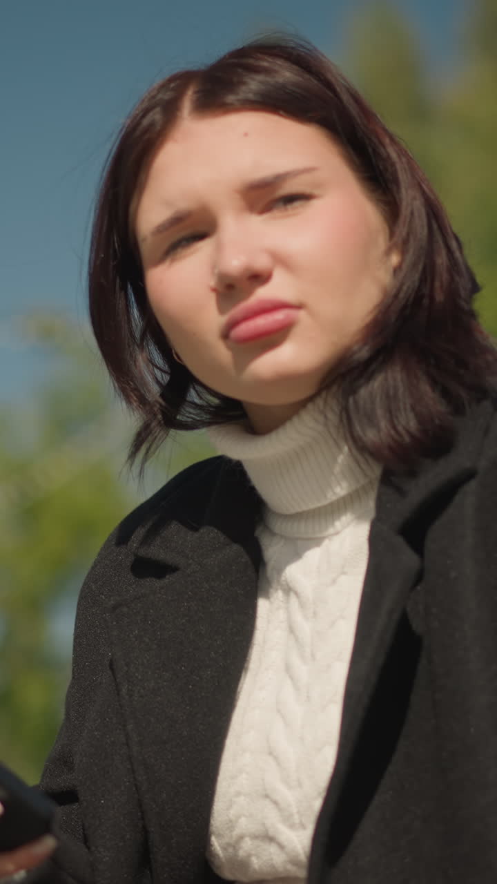 Contemplative woman seated outdoors holding phone, wearing black coat and white sweater, looking away with a thoughtful expression, ring-adorned hand visible, with blurred greenery in background