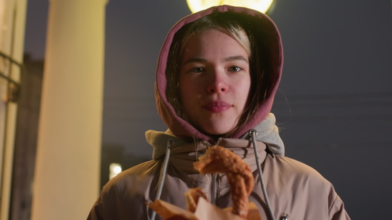 joven de pie al aire libre con pastel en las manos, chaqueta con capucha y cielo nocturno con reloj en el fondo crean un ambiente urbano acogedor