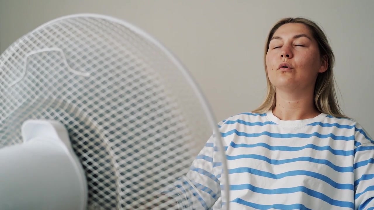 Adulto cansado e superaquecido, com uma blusa listrada, inclina-se em direção a um ventilador elétrico giratório, de olhos fechados, desfrutando do ar fresco durante uma onda de calor em casa