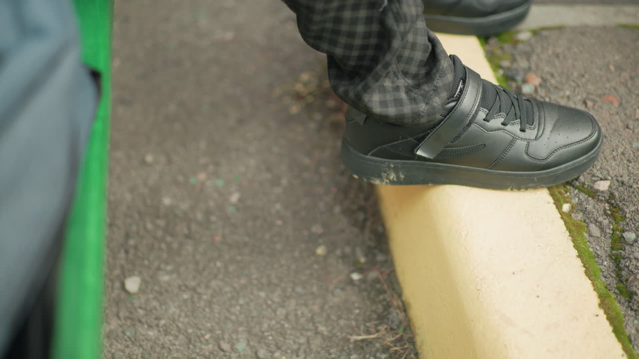 Close up of person legs wearing black sneakers and checkered pants, foot placed on yellow pavement edge outdoors, showing casual street style and natural urban setting