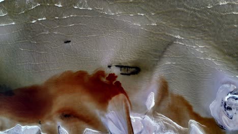 Aerial-footage-of-shipwreck-on-a-sandy-beach-with-turquoise-water,-partially-submerged,-revealing-its-wooden-hull-and-broken-mast,-large-amount-of-oil-on-the-sand