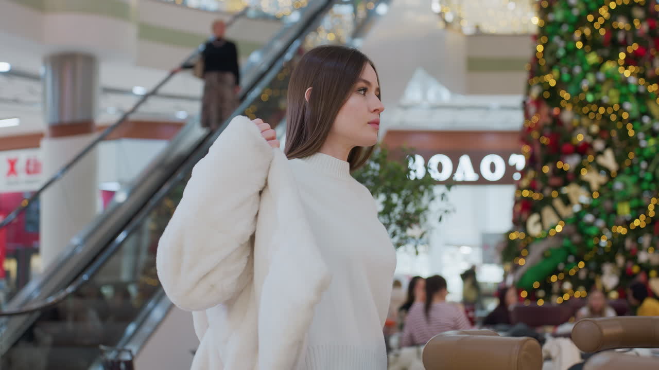Sober woman putting on winter jacket in beautifully decorated shopping mall with large Christmas tree and glowing signage in background