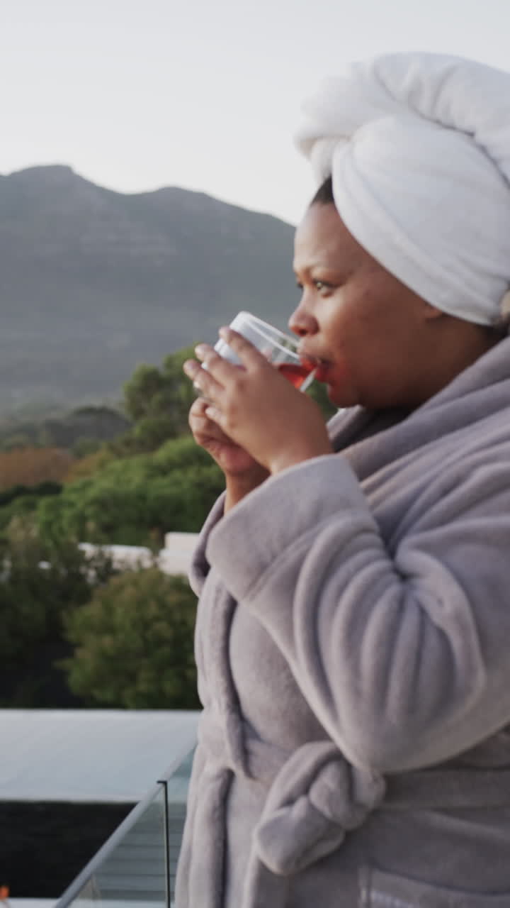 Vertical video of happy plus size african american woman in bathrobe drinking tea on balcony