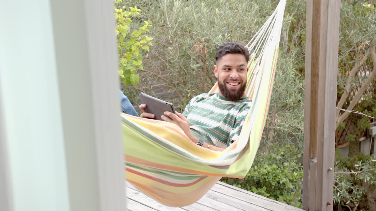 Using tablet and smiling, man relaxing in hammock outdoors