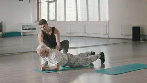 fitness coach helping senior man training plank exercises in sport club