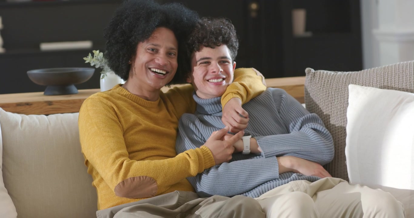 Padre afroamericano e figlio adolescente seduti sul divano, il padre nota lo smartwatch, il figlio si appoggia e sorride