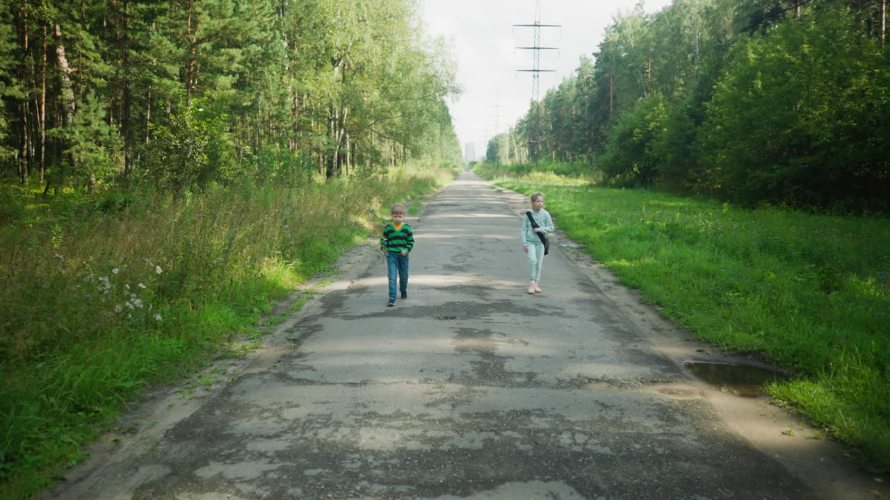 Brother and sister walking apart on tarred road bordered by forest trees and green grass with water puddles reflecting light, enjoying peaceful nature scene in calm outdoor countryside setting