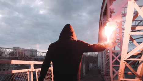 back view of a young man in hoodie and balaclava with red burning signal flare on the road under an old steel frame bridge, slow motion