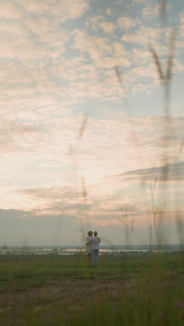 a couple stands on a grassy hill beside a tranquil lake at sunset, gazing at the sky. the man, in a white shirt, hat, and jeans, holds the woman by the waist. she wears a black hat and a white dress