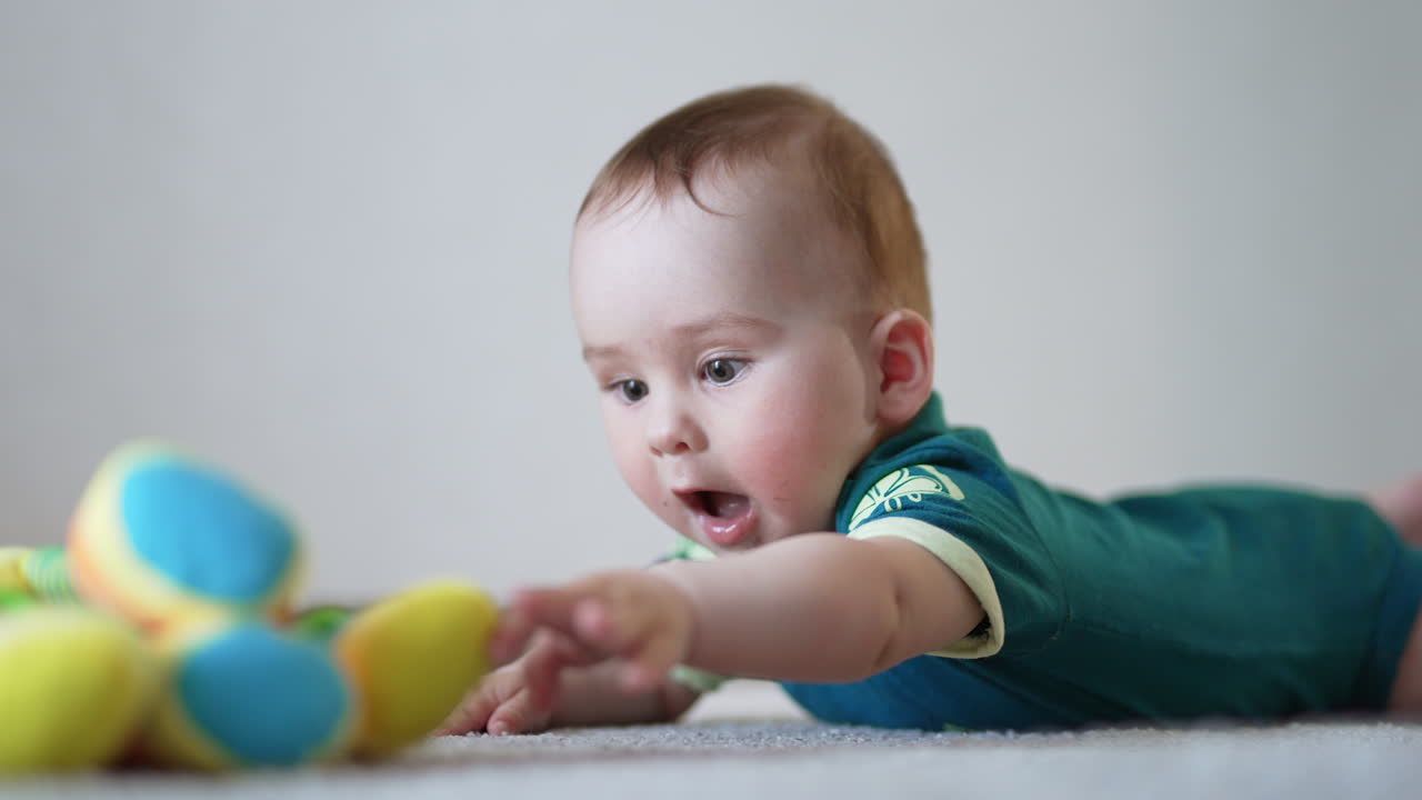 Active baby waving hands lying on the floor. Caucasian child trying to reach a toy in front of him. Light backdrop in blur.