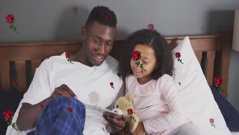 Animation-of-roses-over-happy-african-american-father-and-daughter-reading-book-in-bed