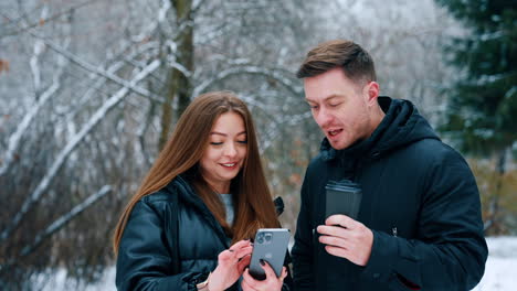 Long-haired brunette showing the smartphone to a man. Caucasian male with cup in hand looks at phone, says something and walks away.