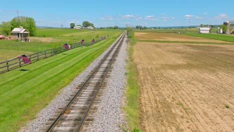 a drone static view of a single empty rail road track that goes thru green farmlands with a fence with american flags on it, on a spring day