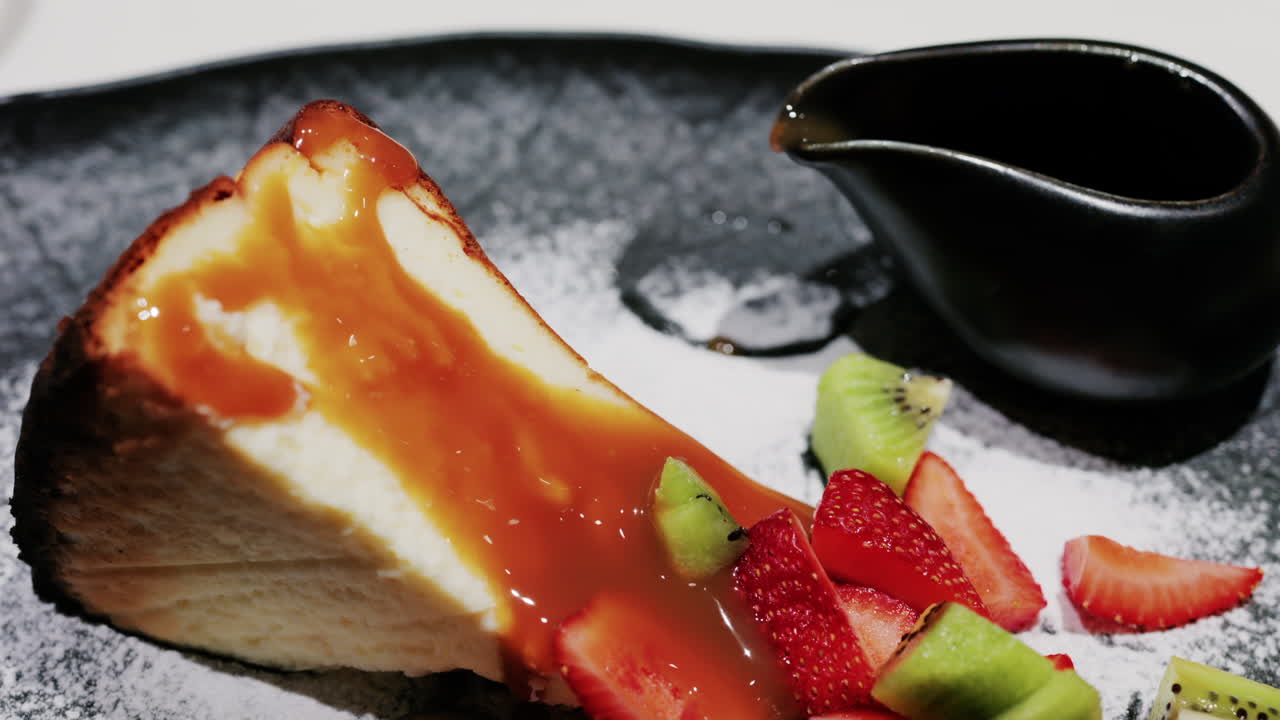 A slice of cheesecake served with caramel sauce fresh strawberries and kiwi, garnished with powdered sugar on a plate at a restaurant
