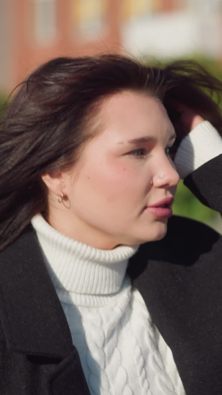 joven con cabello marrón en abrigo negro ajustando su cabello mientras mira a su izquierda, vestida con un suéter de tejer de cable blanco, de pie en una calle urbana soleada