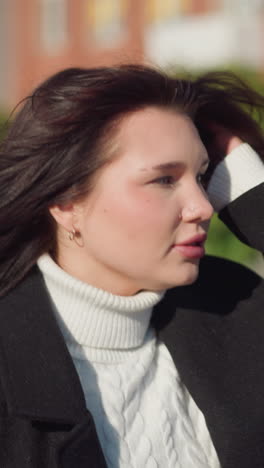 young lady with brown hair in black coat adjusting her hair while looking to her left, dressed in a white cable knit sweater, standing on a sunny urban street