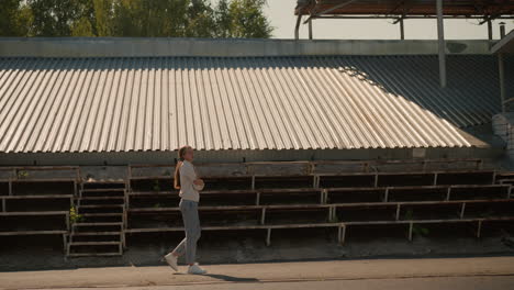 lady in hood and jeans with long hair tied back walking near empty stadium seating on sunny day, hands folded, looking thoughtful and introspective, surrounded by empty bleachers and green trees