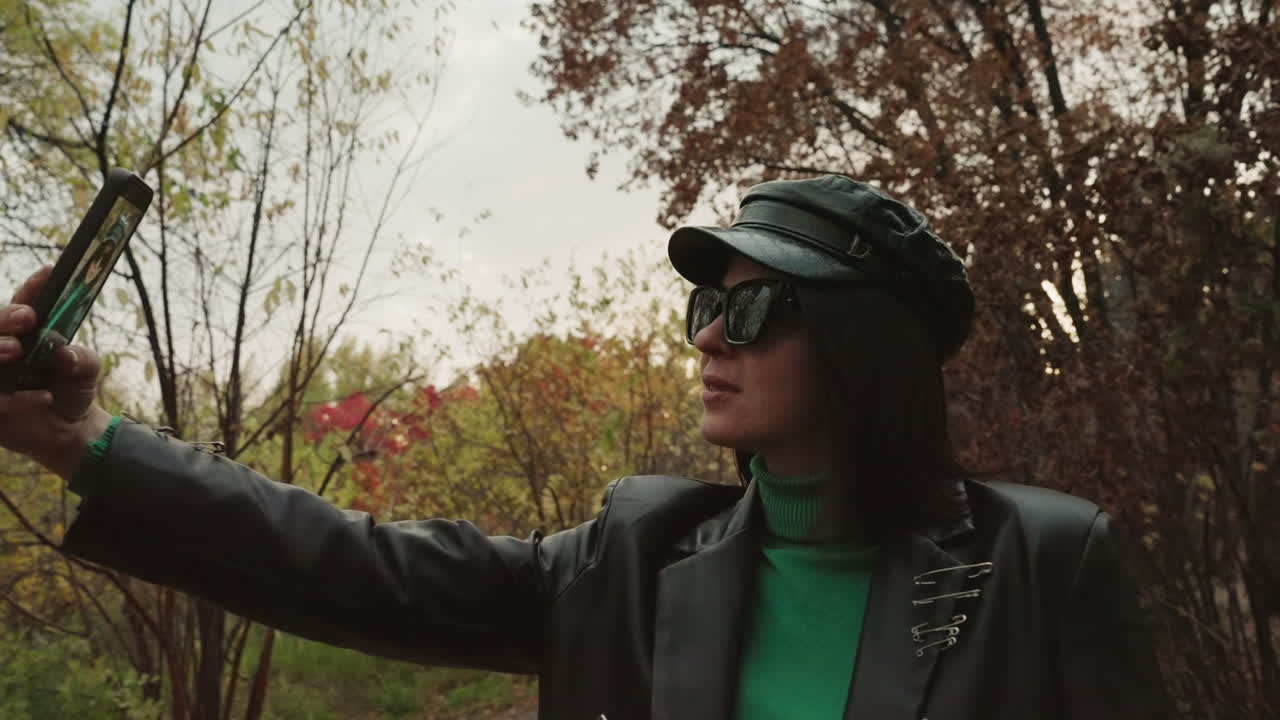 Donna bianca con giacca di pelle che fa un selfie su un sentiero alberato autunnale, tenendo il telefono con il braccio teso, indossa un cappello nero, occhiali da sole oversize, un dolcevita verde e una giacca borchiata, sicura di sé