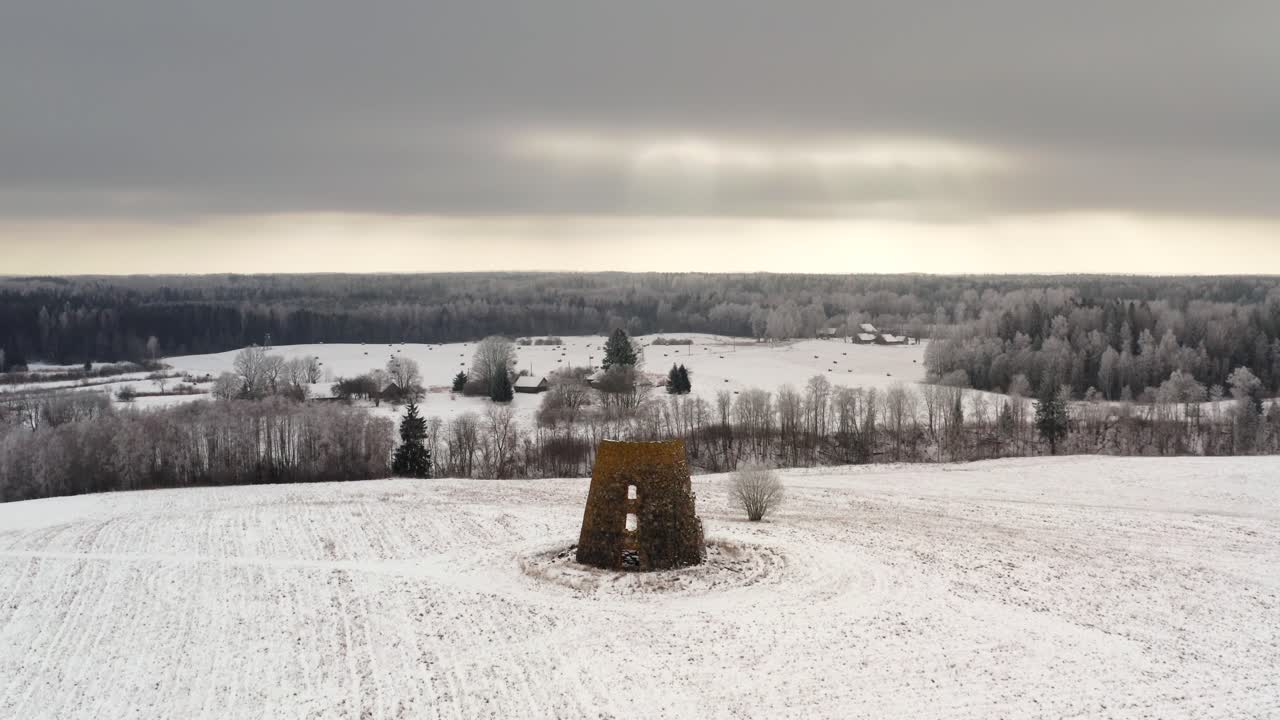 Aerial view of historic windmill ruins on top of a small mountain in countryside. Orbiting drone shot in winter over rural landmark with small village houses in the background. Sunrays through clouds.