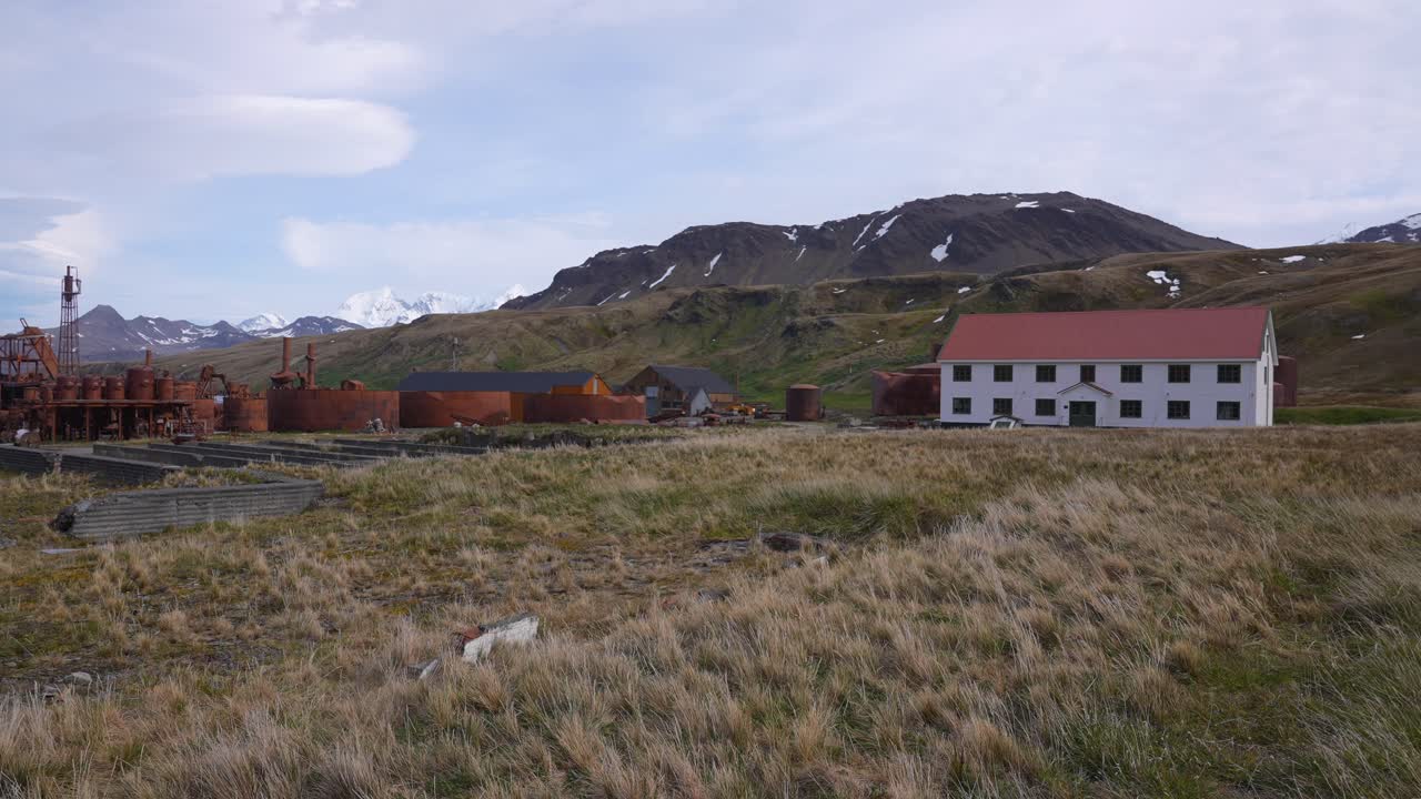 An old industrial site with rusty structures and buildings set against a backdrop of mountains and grassy terrain.