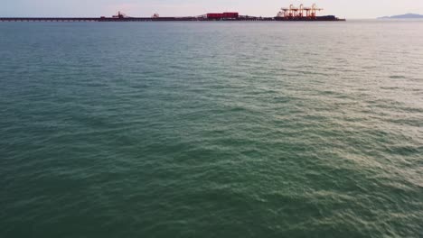 Aerial-drone-flyover-Malaysian-traditional-fishing-boat-tilt-up-reveals-export-wharf-of-brazilian-iron-ore-mining-corporation,-teluk-rubiah-vale-terminal-along-the-horizon-at-dusk