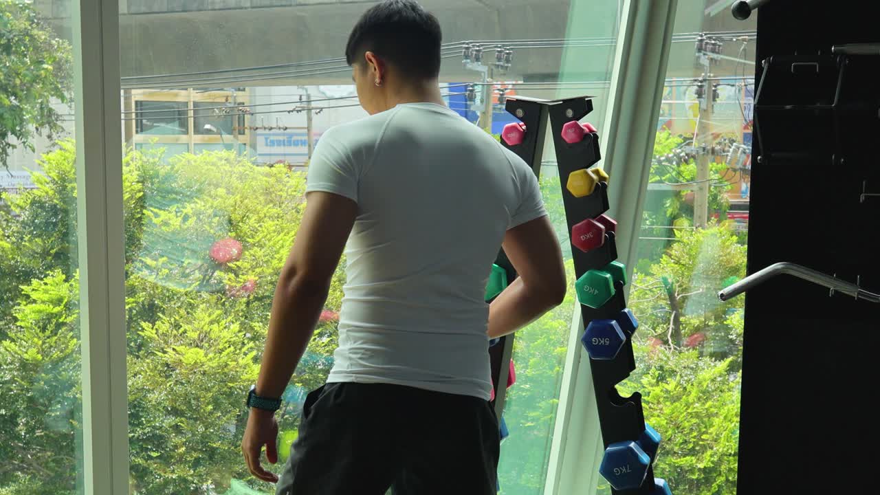 A man in a gym selects dumbbells near a large window with a city view. Bright natural lighting enhances the scene